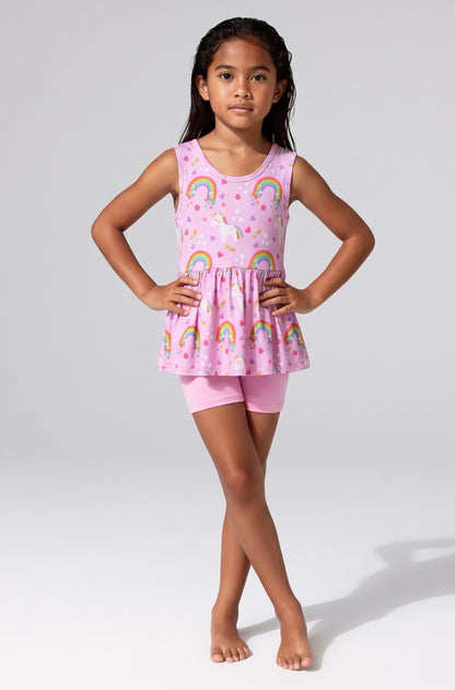 A young girl stands against a light gray studio background wearing a pink sleeveless bamboo viscose peplum tunic top and matching pink shorts. The tunic has an allover rainbow and cloud print on a pink base.