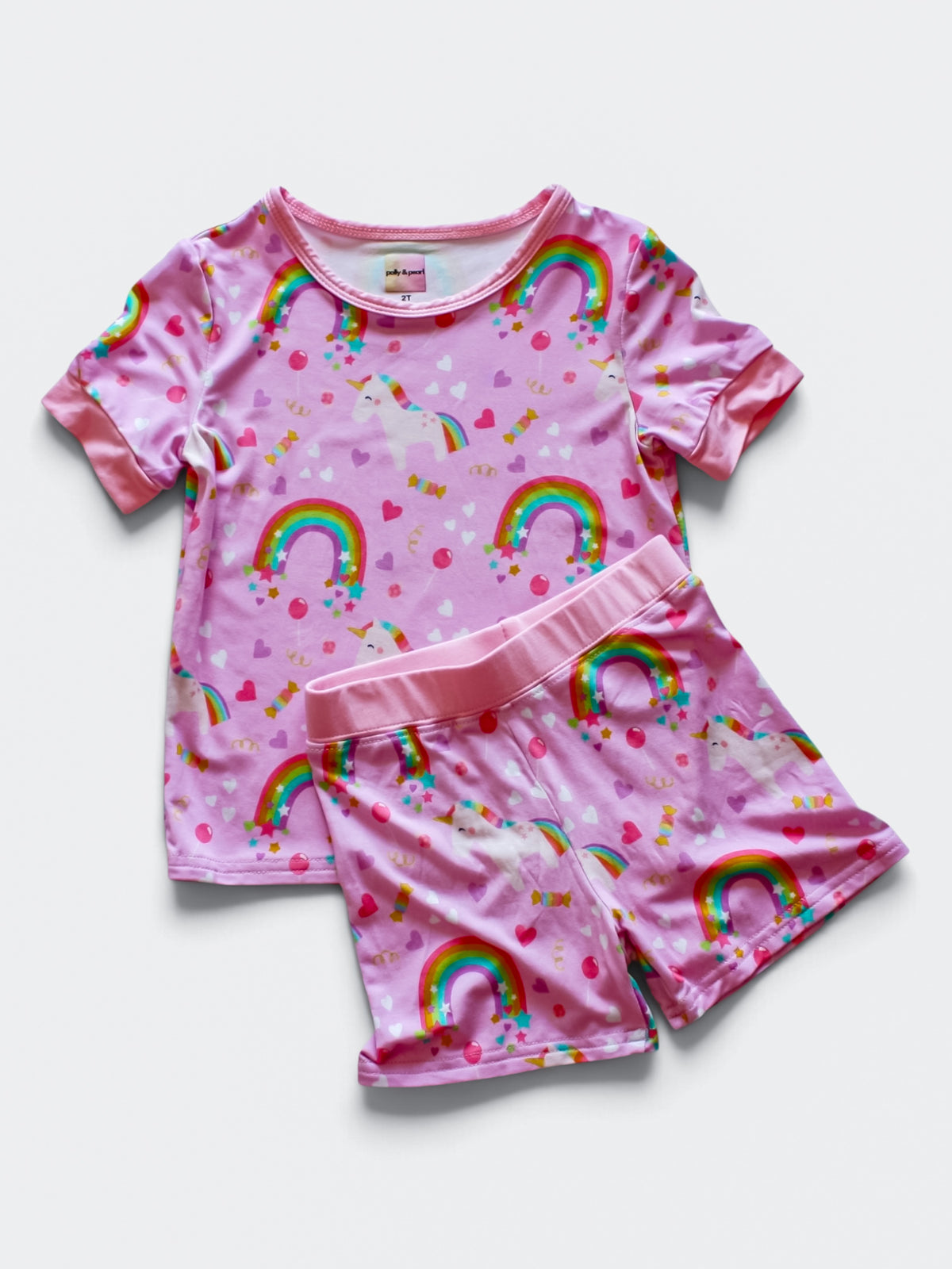 A two-piece kids' bamboo viscose pajama set featuring a short-sleeve top and matching shorts in pink, decorated with a colorful rainbow and unicorn pattern, laid flat on a light gray background.