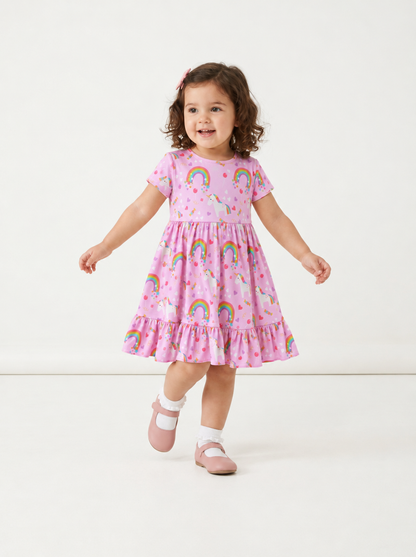A toddler girl wearing a pink bamboo viscose short-sleeve dress with a rainbow and unicorn print, walking on a seamless white studio background.