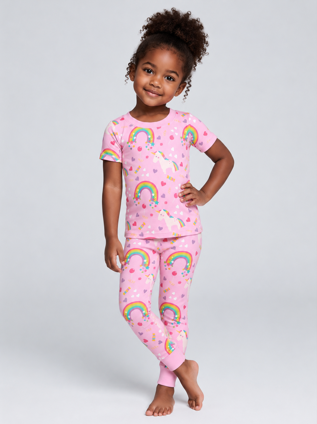 A young girl with curly hair stands barefoot against a light gray studio background, wearing a pink bamboo viscose short-sleeve pajama top and matching pajama pants with a rainbow and unicorn print.