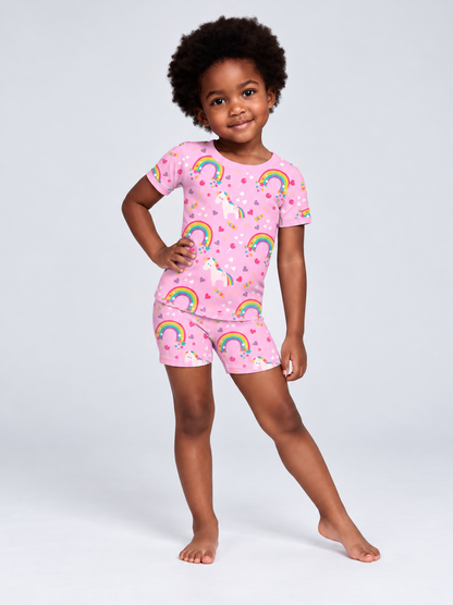 A young child modeling a pink bamboo viscose  two-piece pajama set with a colorful rainbow and unicorn pattern, standing against a light gray studio background.
