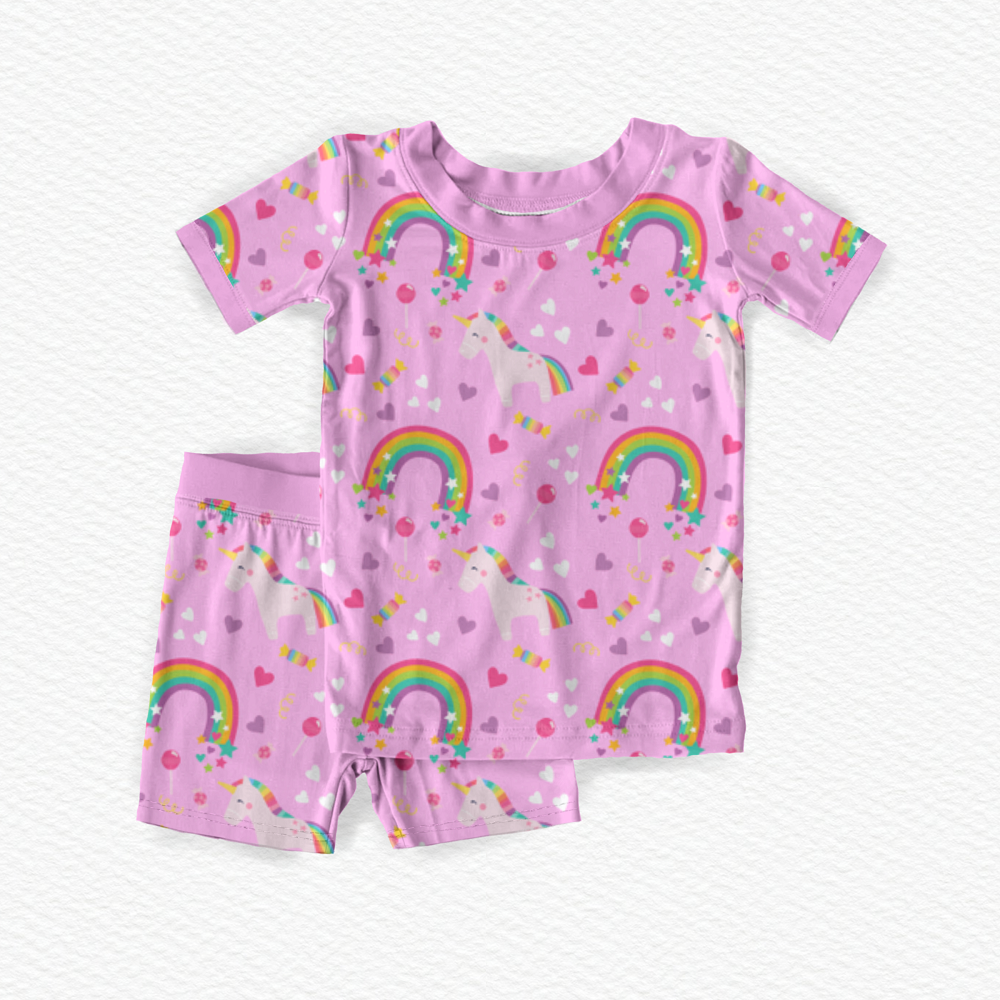 A two-piece children's bamboo viscose pajama set featuring a short-sleeve top and matching shorts in pink, decorated with a colorful print of unicorns, rainbows, hearts, and stars.